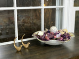 Two small gold bird figurines next to a white ceramic bowl filled with dried purple and beige potpourri on a dark wooden windowsill with white window frames.