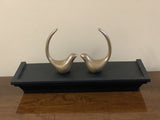 Two smooth bronze bird figurines facing each other with curved raised tails, displayed on a rectangular black wooden tray atop a polished wooden surface.