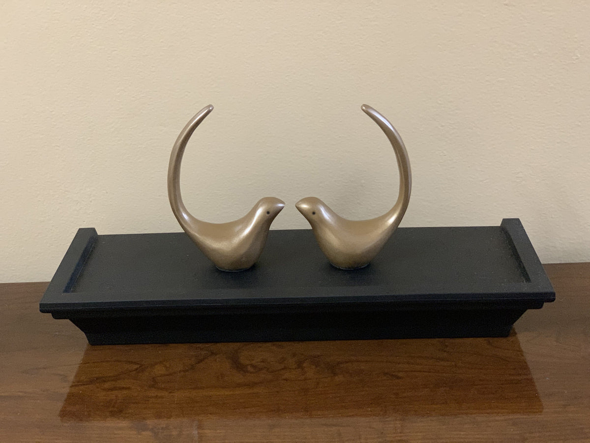 Two smooth bronze bird figurines facing each other with curved raised tails, displayed on a rectangular black wooden tray atop a polished wooden surface.
