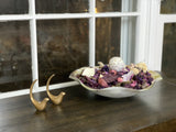 Two small brass bird figurines with curved tails next to a decorative shallow bowl filled with purple potpourri, displayed on a dark wooden surface by a window.