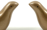 Close-up of two golden-brown bird figurines facing each other with smooth, metallic textured surfaces and small black eyes on a white background.