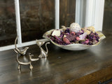 Three small silver animal figurines shaped like a dog, cat, and bird displayed on a dark wooden surface near a window, with a metal bowl of dried flowers behind them.