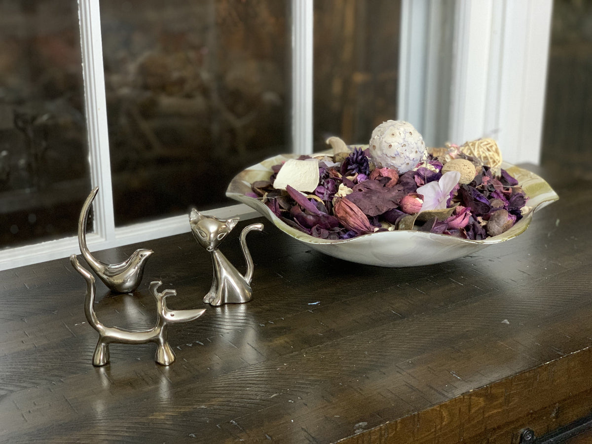 Three small silver animal figurines shaped like a dog, cat, and bird displayed on a dark wooden surface near a window, with a metal bowl of dried flowers behind them.