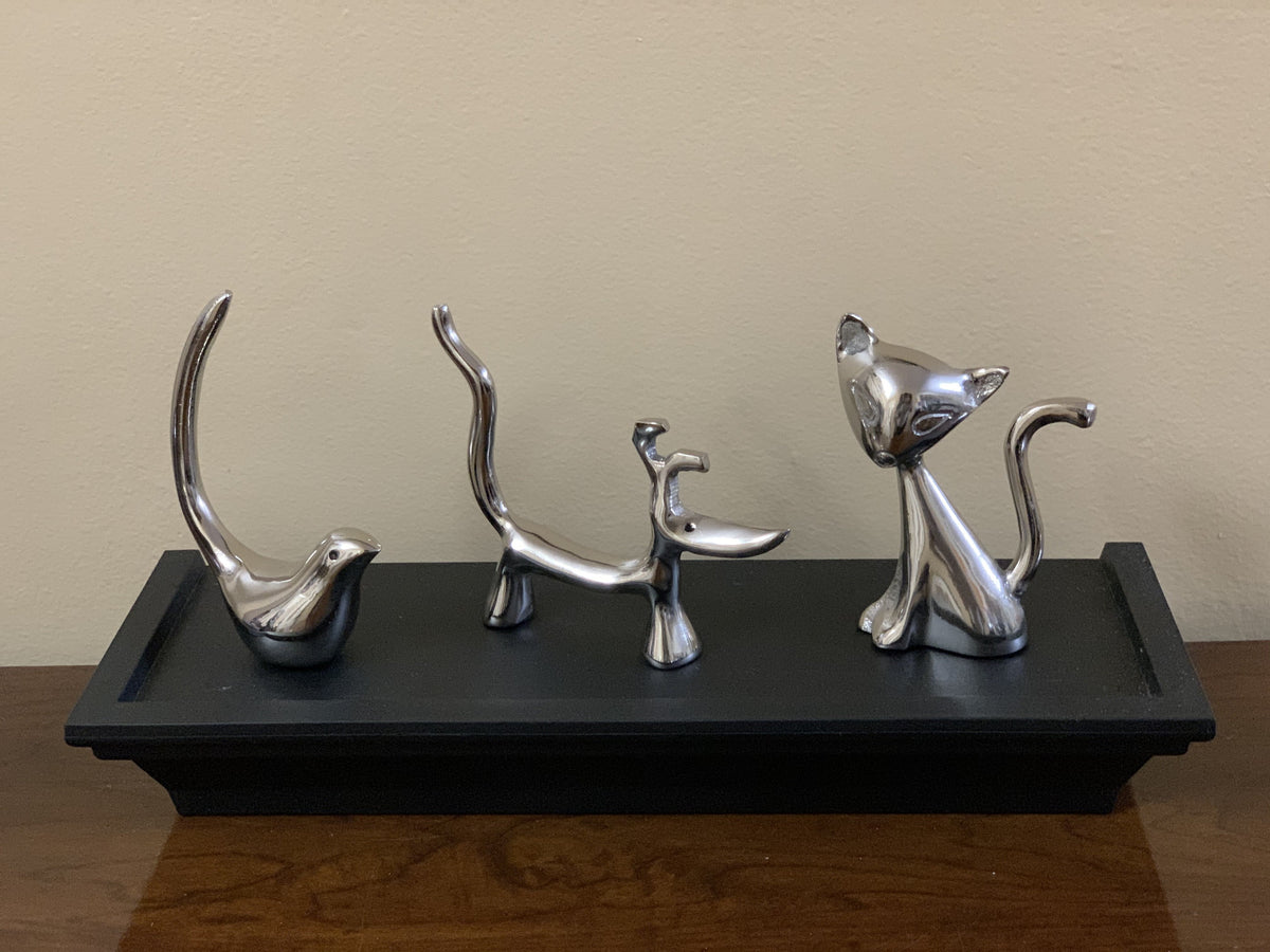 Set of four sleek silver ring holders shaped like a bird, dog, cat, and standing figure, displayed on a black rectangular tray on a wooden surface.