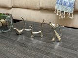 Three small silver ring holders shaped like a bird, a dog with tail up, and a cat with big eyes, displayed on a gray wooden surface with a beige couch and blue patterned cloth in background.