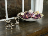 Three silver animal-shaped ring holders—a bird, cat, and dachshund—displayed next to a ceramic dish filled with dried purple and beige potpourri on a dark wood surface.