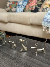 Three silver ring holder figurines shaped like a sitting dog, a cat with an upright tail, and a dachshund, arranged on a dark wooden table with a blurred beige sofa and colorful cushions in the background.