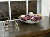 Silver-toned metal figurines of a dog, cat, and elephant arranged on a wooden surface beside a decorative bowl filled with purple and beige dried petals and spheres.