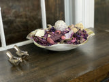 Silver rustic bird figurines next to a decorative bowl filled with dried purple and beige potpourri on a wooden surface near a window.