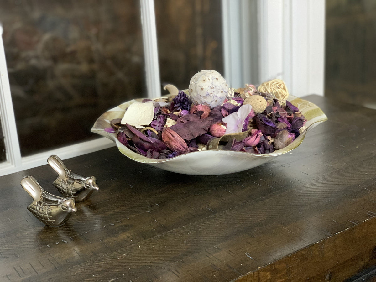 Silver rustic bird figurines next to a decorative bowl filled with dried purple and beige potpourri on a wooden surface near a window.
