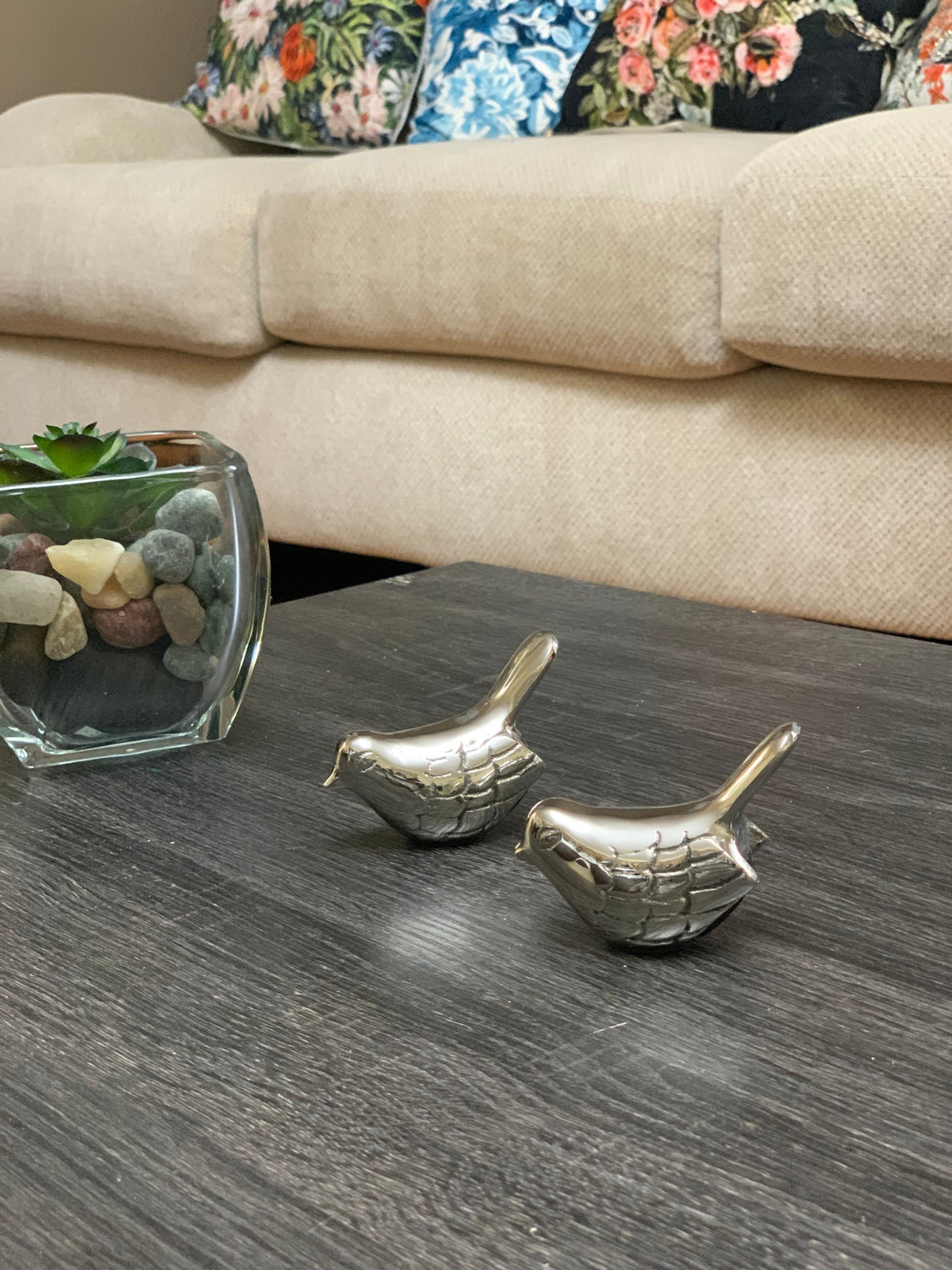 Two small silver bird figurines with engraved feather details on a dark wooden table, next to a glass container with decorative stones and a succulent plant.