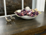 Silver bowl filled with dried purple and pink petals and decorative balls on a wooden surface beside two small silver bird figurines, near a window.