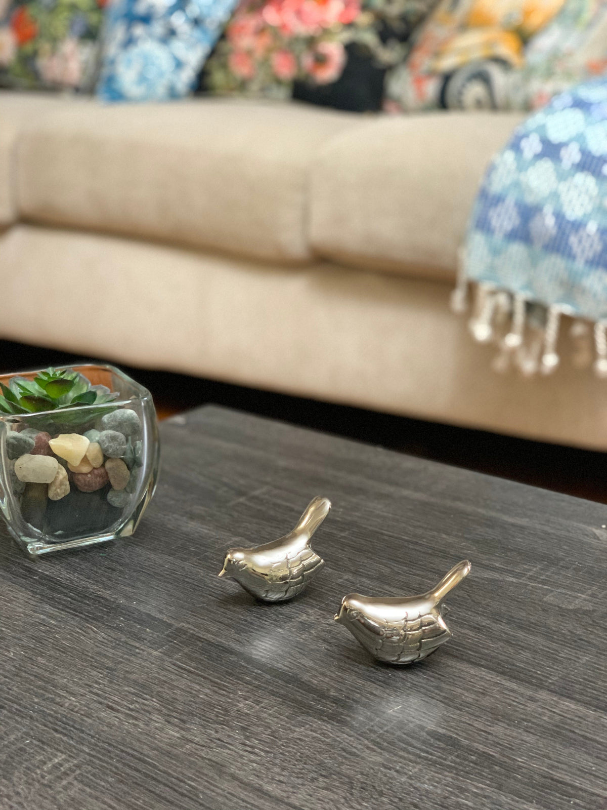 Two small silver bird figurines with a rustic finish placed on a dark wooden table near a glass planter with succulent and pebbles.