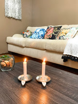 Pair of silver taper candlestick holders with lit white candles on dark wood coffee table, beige sofa with colorful floral pillows and a fringed throw in background.