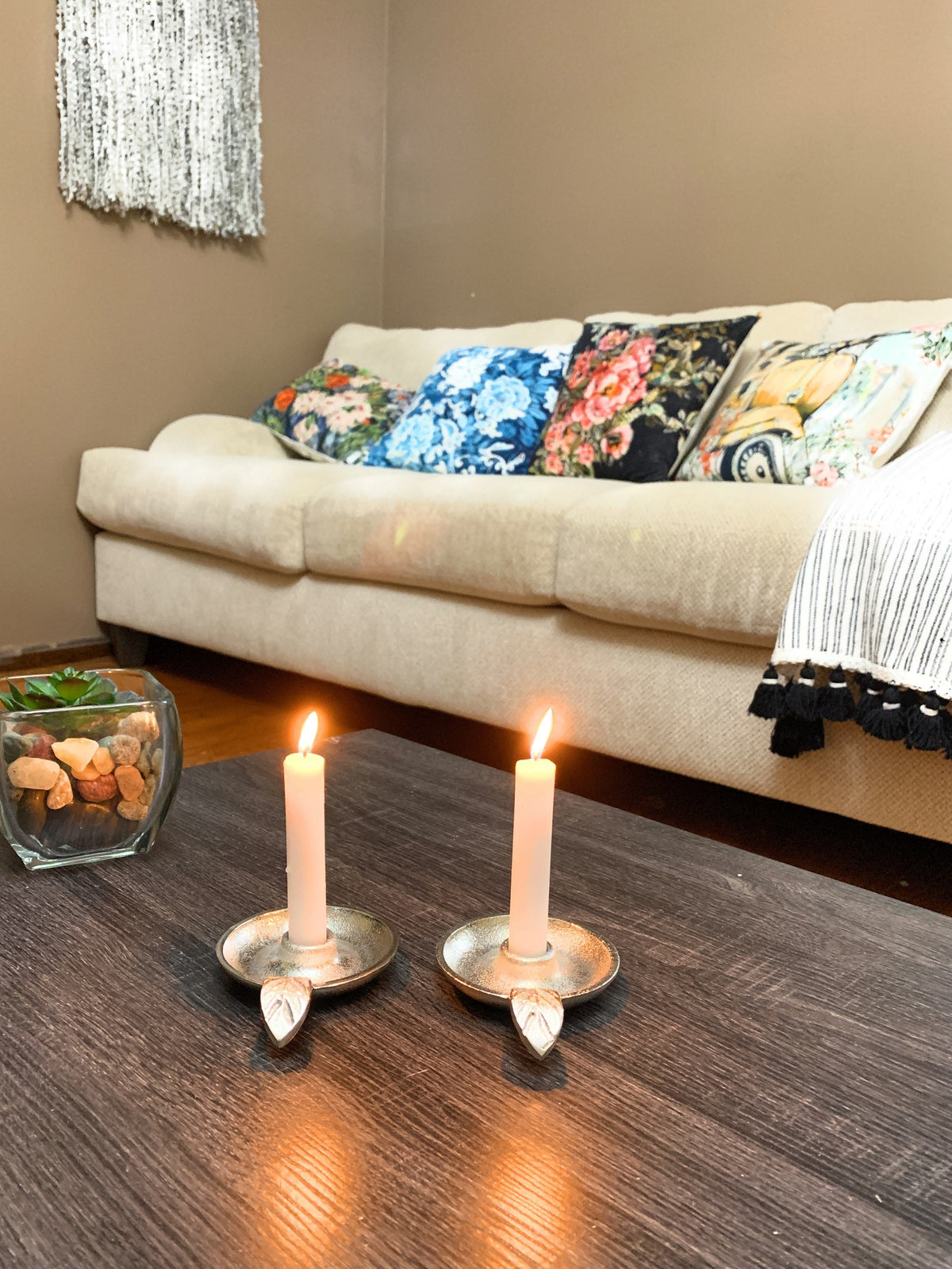 Pair of silver taper candlestick holders with lit white candles on dark wood coffee table, beige sofa with colorful floral pillows and a fringed throw in background.