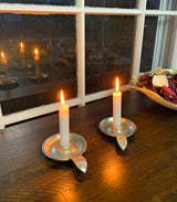 Two silver taper candlestick holders with leaf-shaped handles, each holding a lit white candle, placed on a dark wooden surface near a window reflecting their flames.