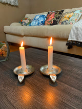 Two lit white taper candles in silver candlestick holders with leaf-shaped handles on a dark wood table, in front of a beige sofa with colorful floral cushions.