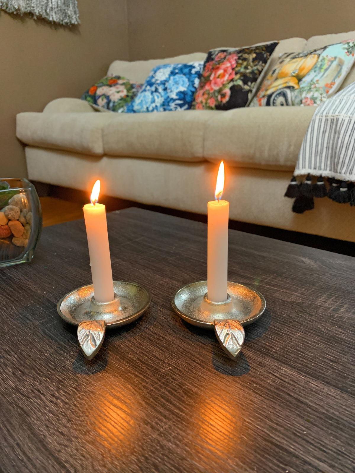 Two lit white taper candles in silver candlestick holders with leaf-shaped handles on a dark wood table, in front of a beige sofa with colorful floral cushions.