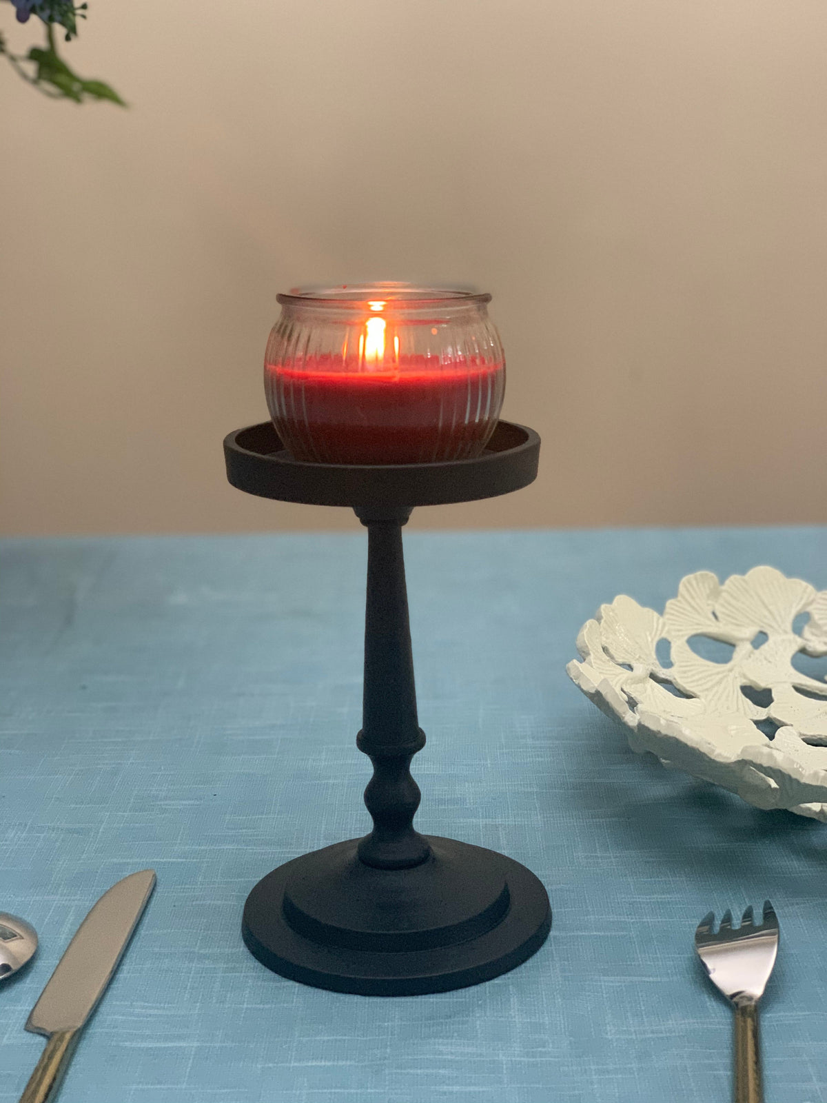 Matte black metal pillar candle holder with a round base and tapered stem, holding a lit red glass jar candle on a blue tablecloth.