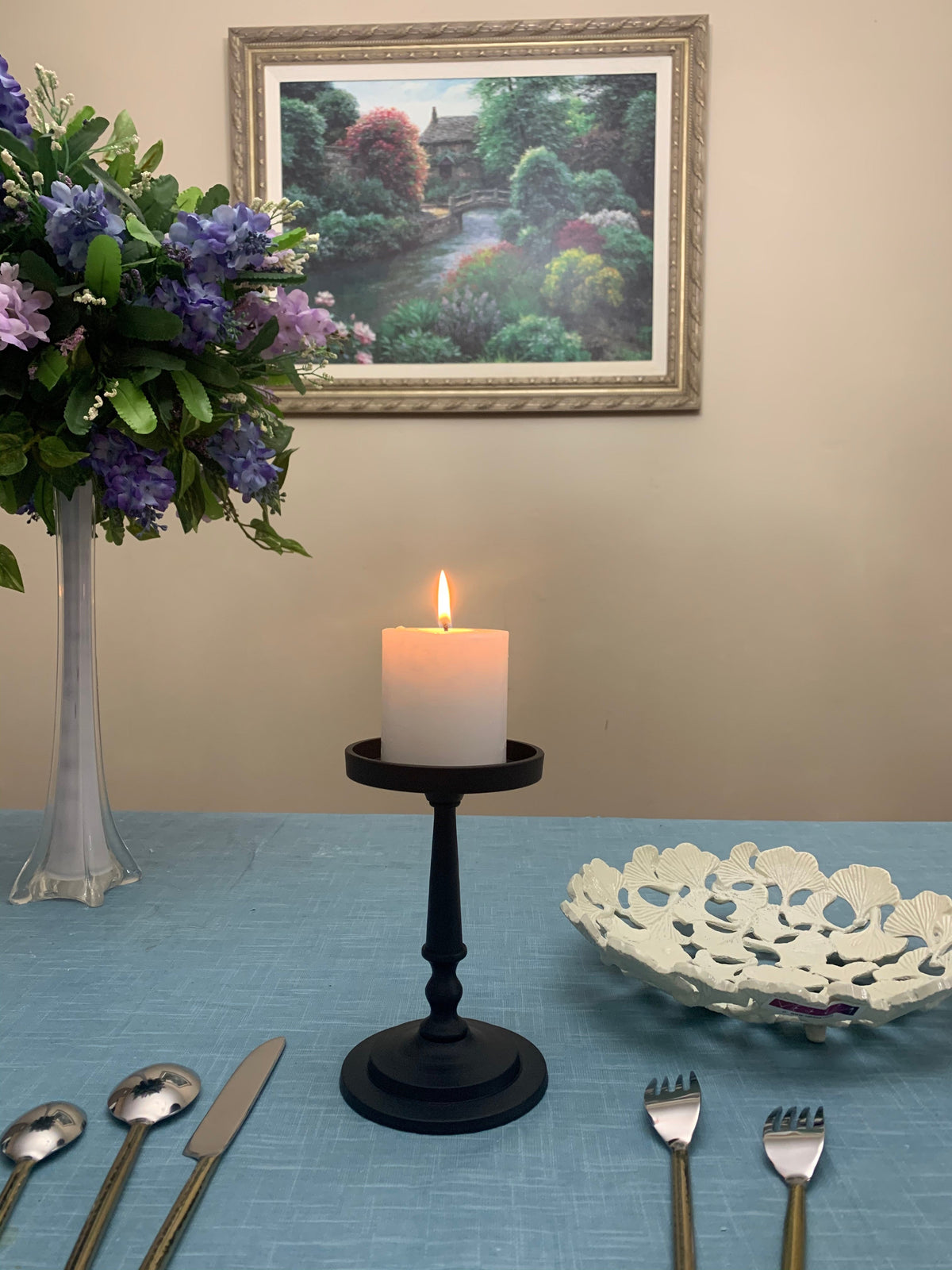 Matte black pillar candle holder with lit white pillar candle on a blue tablecloth, flanked by a flower vase with purple blooms and a decorative white bowl, under a framed landscape painting.