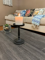 Matte black metal pillar candle holder with a lit white candle on a dark wood coffee table, with a beige couch and colorful floral pillows in the background.