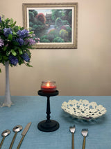 Matte black Vibhsa pillar candle holder with a lit red candle on blue tablecloth, accompanied by a white floral-patterned bowl and silver cutlery, with floral painting background.