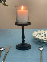 Matte black metal pillar candle holder with a glass hurricane shade holding a lit white candle, placed on a blue textured tablecloth.