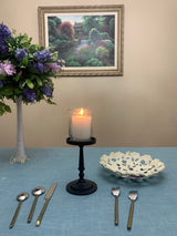 Matte black pillar candle holder with a lit white candle on a blue tablecloth, flanked by a floral arrangement in a tall clear vase and a white decorative bowl with leaf cutouts.