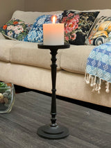 Tall matte black metal candle holder with turned spindle design holding a lit white pillar candle, placed on a dark wood coffee table in front of a beige sofa with floral cushions.