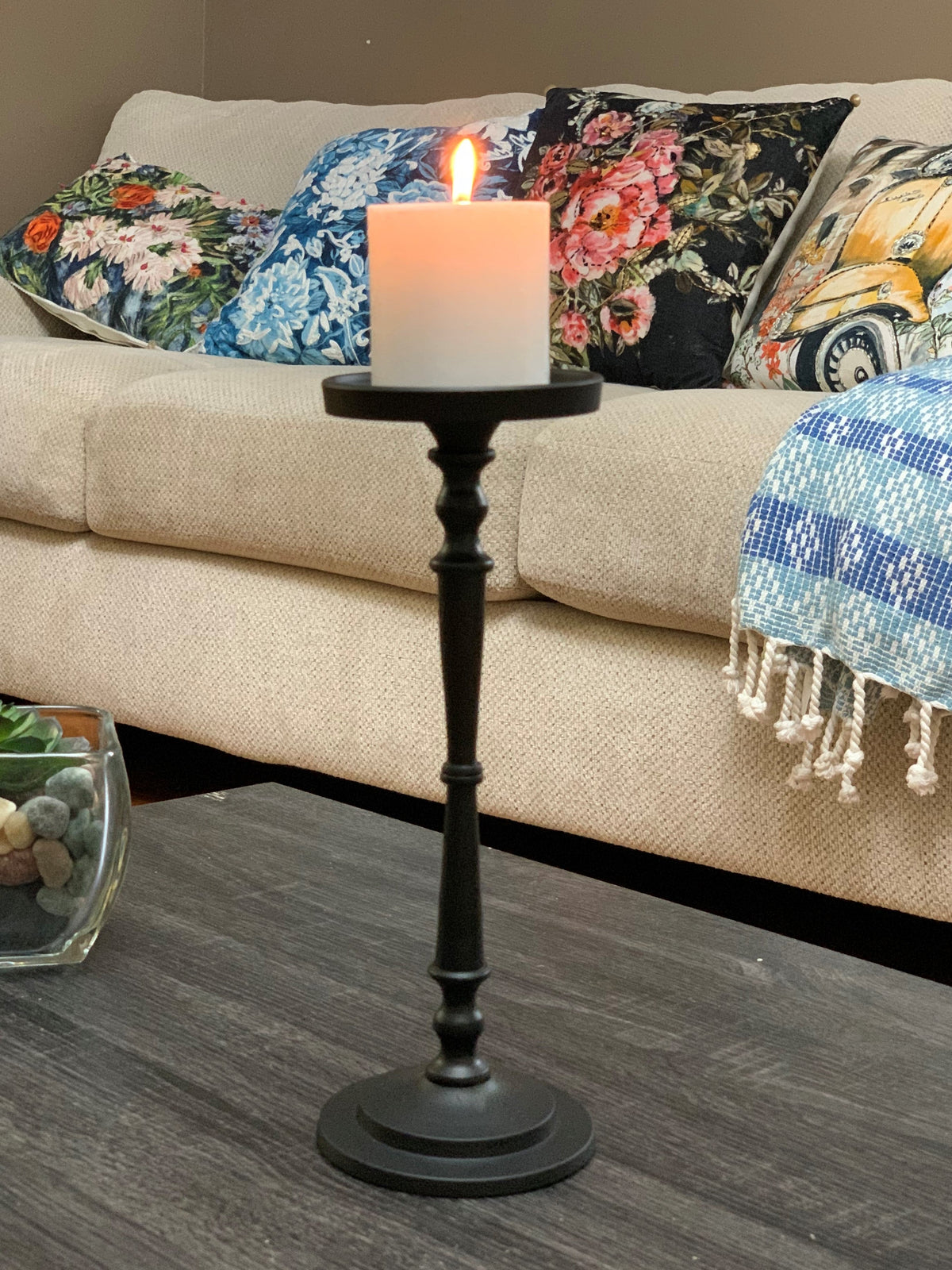 Tall matte black metal candle holder with turned spindle design holding a lit white pillar candle, placed on a dark wood coffee table in front of a beige sofa with floral cushions.