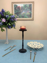 Tall matte black candle holder with a lit red candle, placed on a blue tablecloth with a white decorative bowl, gold-handled forks and spoons, and a vase of purple flowers nearby.