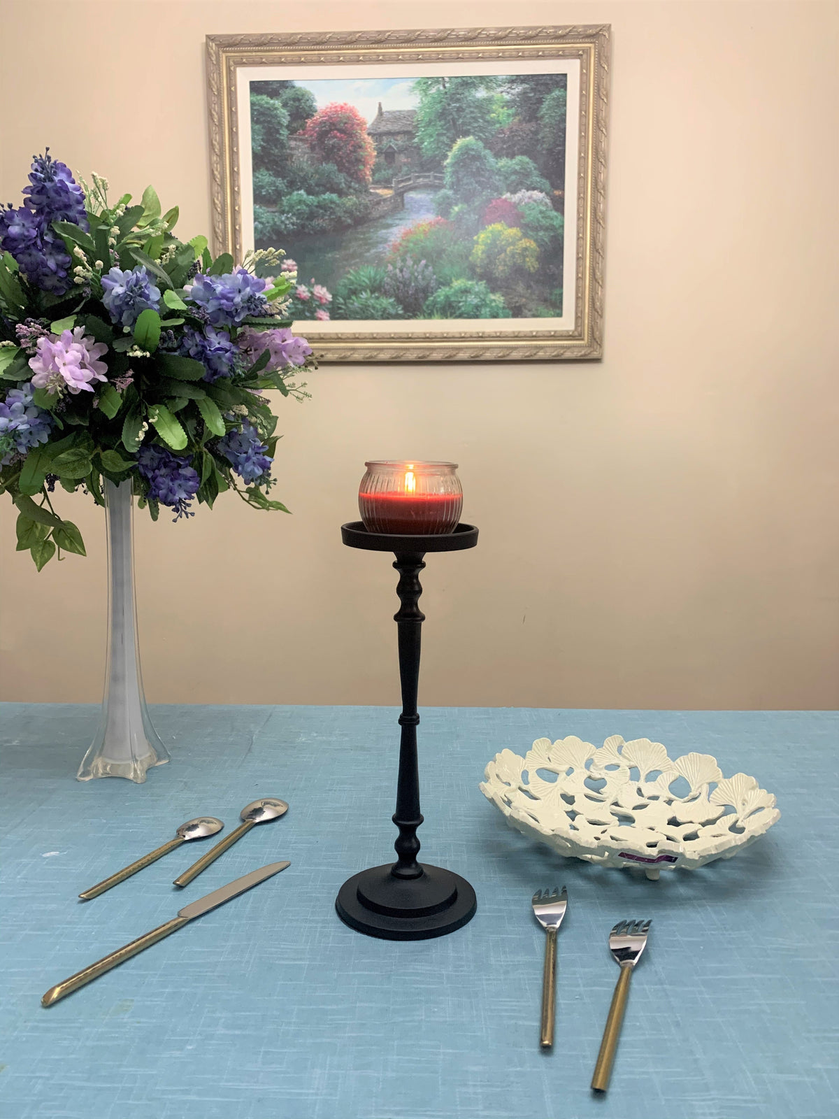 Tall matte black candle holder with a lit red candle, placed on a blue tablecloth with a white decorative bowl, gold-handled forks and spoons, and a vase of purple flowers nearby.