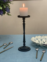 Tall matte black pillar candle holder with a lit white pillar candle, placed on a light blue textured surface with utensils and a decorative white dish nearby.