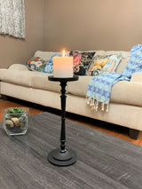 Tall matte black pillar candle holder with a lit white candle on a dark wood coffee table, beige sofa with colorful patterned cushions and a blue throw in background.