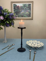 Matte black tall candle holder with a lit white pillar candle on a blue cloth-covered table, surrounded by a decorative white bowl, silver cutlery, and a flower arrangement against a beige wall.