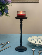 Tall matte black candle holder with a slender, ornate stem supporting a lit red candle in a round glass container, placed on a light blue tablecloth.