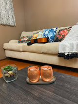 Two hammered copper Moscow Mule mugs filled with ice on a small oval tray on a dark wood table, with a beige couch and colorful floral pillows in the background.