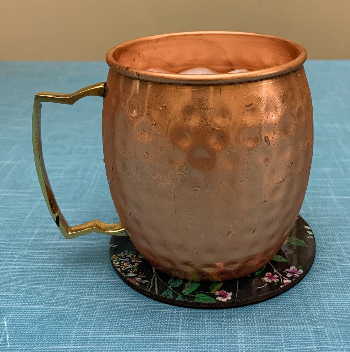 Copper Moscow Mule mug with hammered texture and a brass-colored handle, placed on a floral-patterned round coaster on a blue textured surface.
