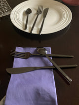 Set of stainless steel dinner knife, fork, and teaspoon arranged on a white plate and on a purple cloth, all placed on a dark wooden table.