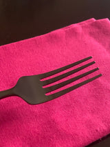 Matte black stainless steel dinner fork with four tines resting on a bright magenta cloth against a dark background.