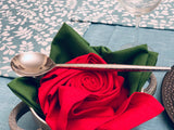 Polished silver table spoon with a textured handle, resting diagonally on a green and red cloth folded like a flower on a patterned blue tablecloth.