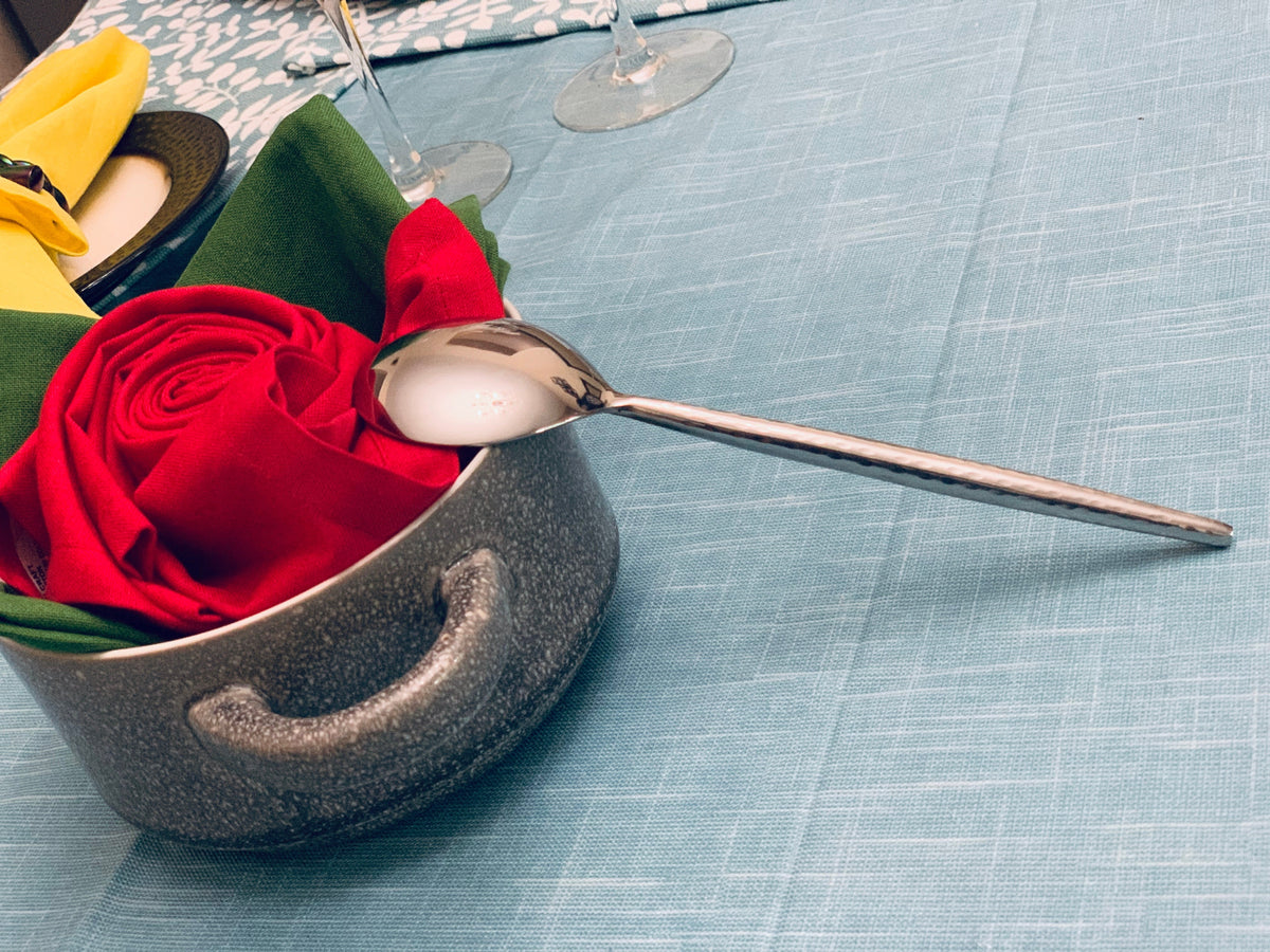 Single polished silver table spoon resting on blue textured fabric, beside a ceramic pot with folded cloths resembling a red rose and green leaves.