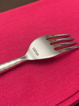Close-up of a polished stainless steel fork engraved with 'Vibhasa' on the handle, resting diagonally on a textured bright pink fabric background.