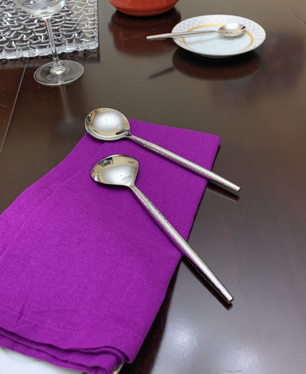 Two polished silver spoons with round bowls resting on a folded deep purple napkin on a dark wood table, with a glass and small white plate in the background.