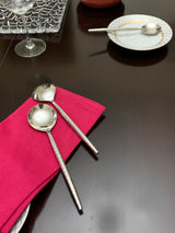 Two polished silver serving spoons with hammered handles resting on a magenta cloth on a dark wooden table, with a small spoon on a patterned plate nearby.