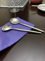 Two shiny stainless steel spoons with hammered handles resting on a folded purple napkin on a dark wood table near a wine glass and white plate.