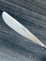 Close-up of a silver stainless steel serrated knife with the brand name Vibhasa engraved on the blade, resting on a dark textured wooden surface.