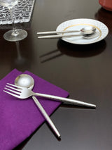 Hammered stainless steel fork and knife crossed on a purple napkin, with matching spoon and chopsticks resting on a white patterned plate on a dark reflective table.