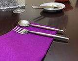 Polished stainless steel spoon and fork with hammered texture handles resting on a vibrant purple napkin on a dark reflective table.