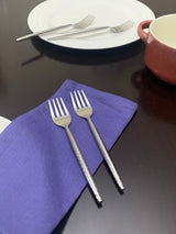 Two small stainless steel appetizer forks with textured handles resting on a folded purple cloth, with a white plate and fork in the background on a dark surface.